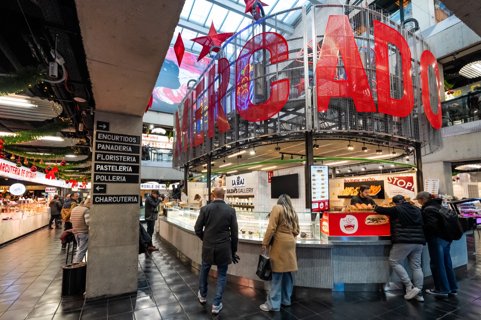 Inside a bustling Madrid food market, shoppers at a bocata and charcutería counter beneath a big red MERCADO sign