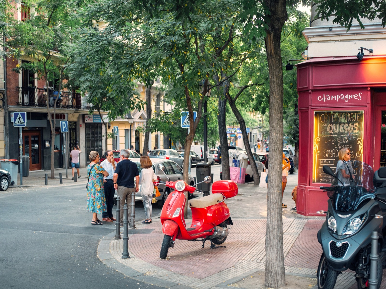A leafy Madrid corner with a red Vespa parked outside a champagne and cheese bar, neighbors chatting on the pavement