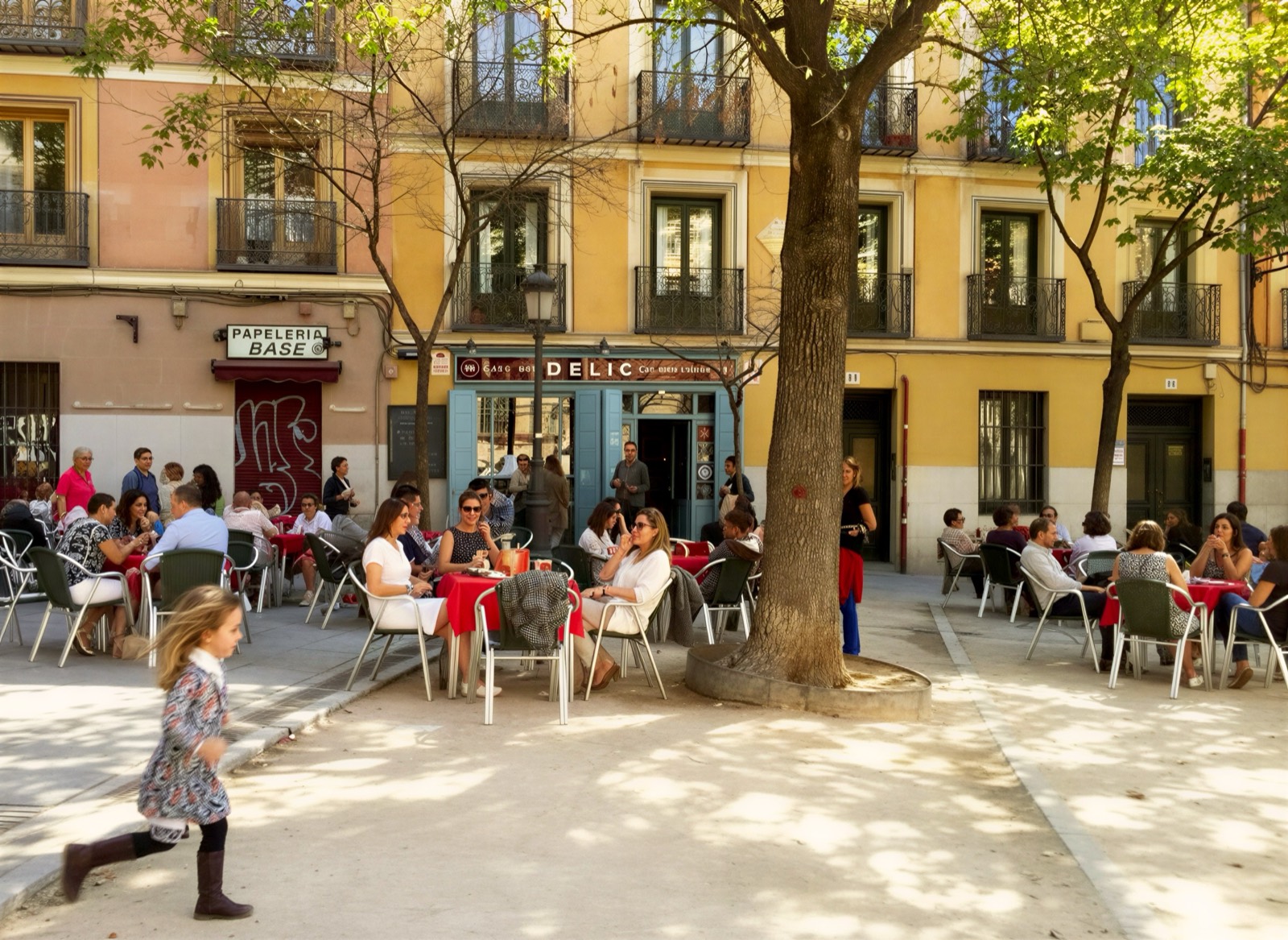 A child running across a sunny Madrid plaza past the busy terraces of Café Delic in La Latina